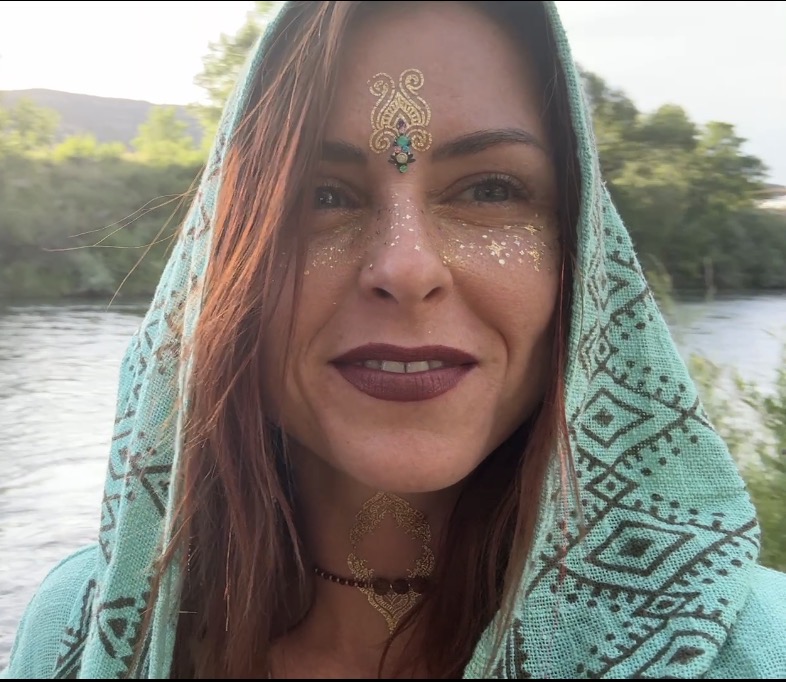 Shiffy in a teal hood with gold festival face art by the river